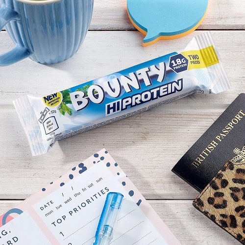 Bounty Hi-Protein Bar 52g protein snack bar with 18g protein, coconut and chocolate flavour, shown in its white and blue packaging on a desk with accessories.