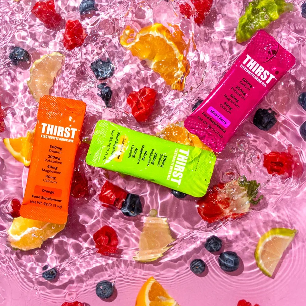 Thirst Electrolyte Drink Mix single-serve sachets in assorted flavours, including orange, mixed berry, and lime, shown with fruit slices and berries in water. Hydration supplement with sodium, potassium, magnesium, and calcium.