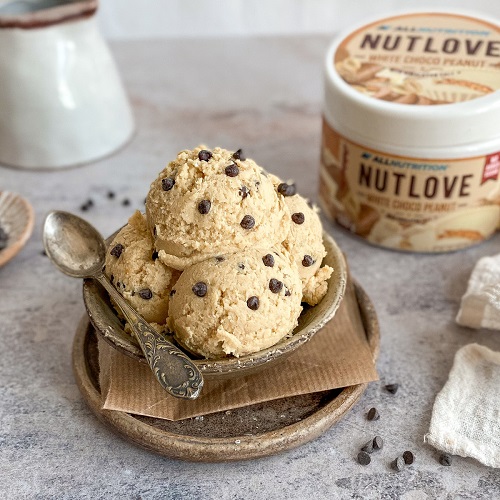 Allnutrition Nutlove White Choco Peanut 500g creamy peanut spread with white chocolate, shown in a bowl with chocolate chips and the product tub in the background. Light beige color dominates the scene.